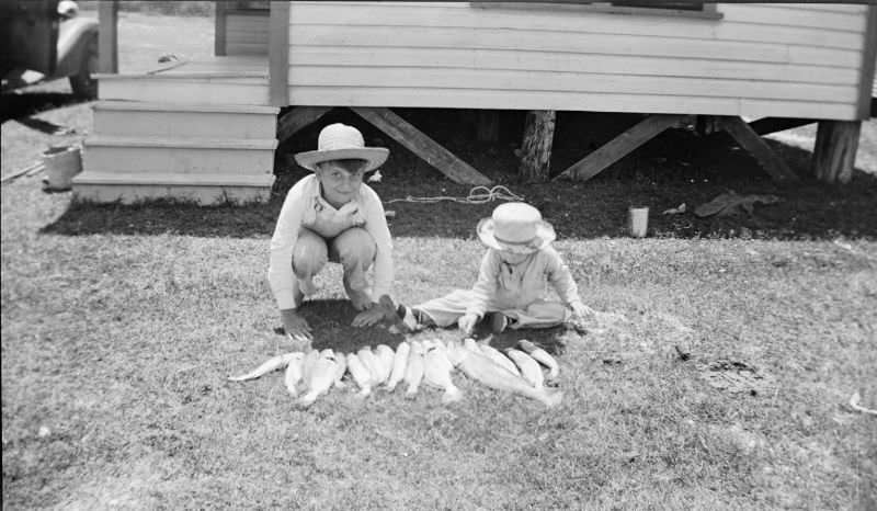 #34 Two boys kneeling behind a line of fish in the grass