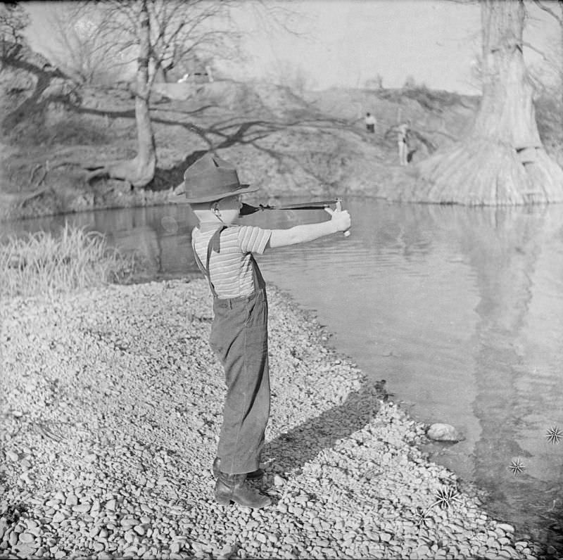 #8 Boy playing with a toy slingshot, standing on a river bank