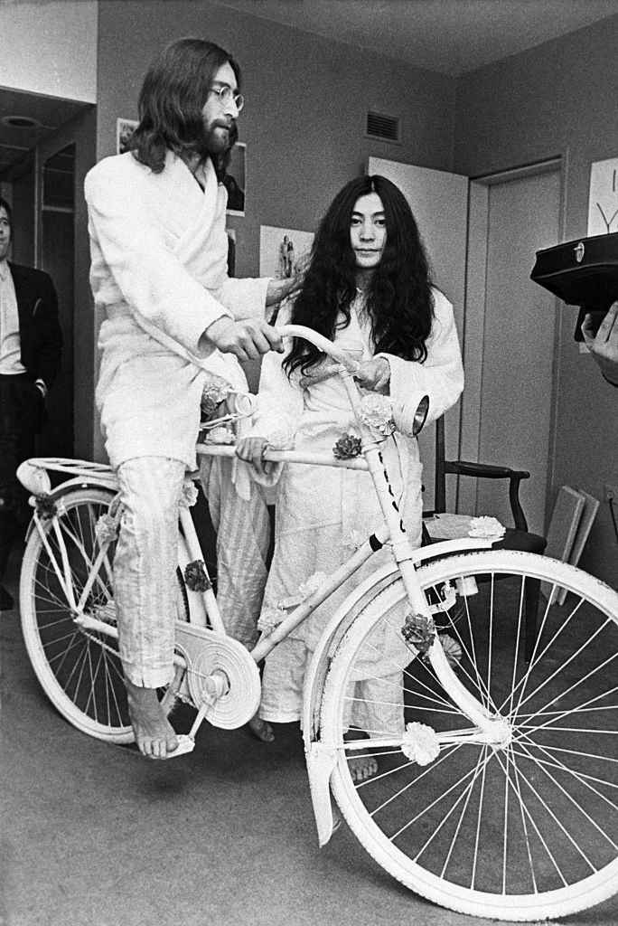 #16 Beatle John Lennon tries out a bicycle he received as a gift while his new wife, Yoko Ono, stands by in Amsterdam, 1969.