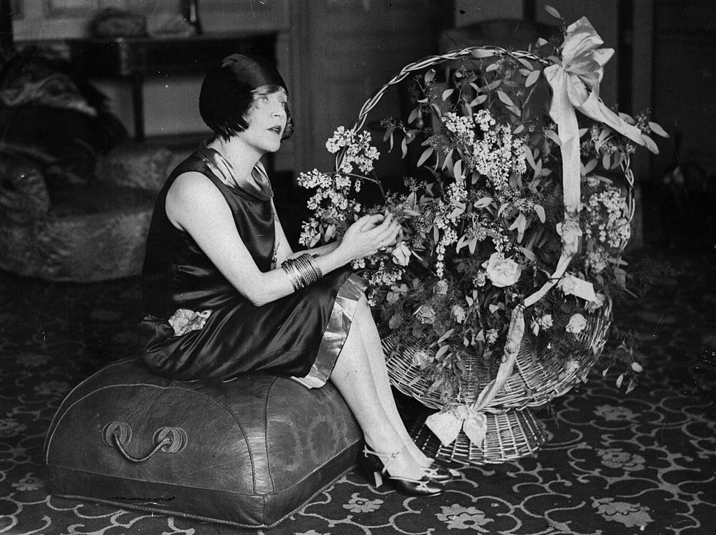 #26 Mae Murray seated on a pouffe with a large basket of flowers, 1925.