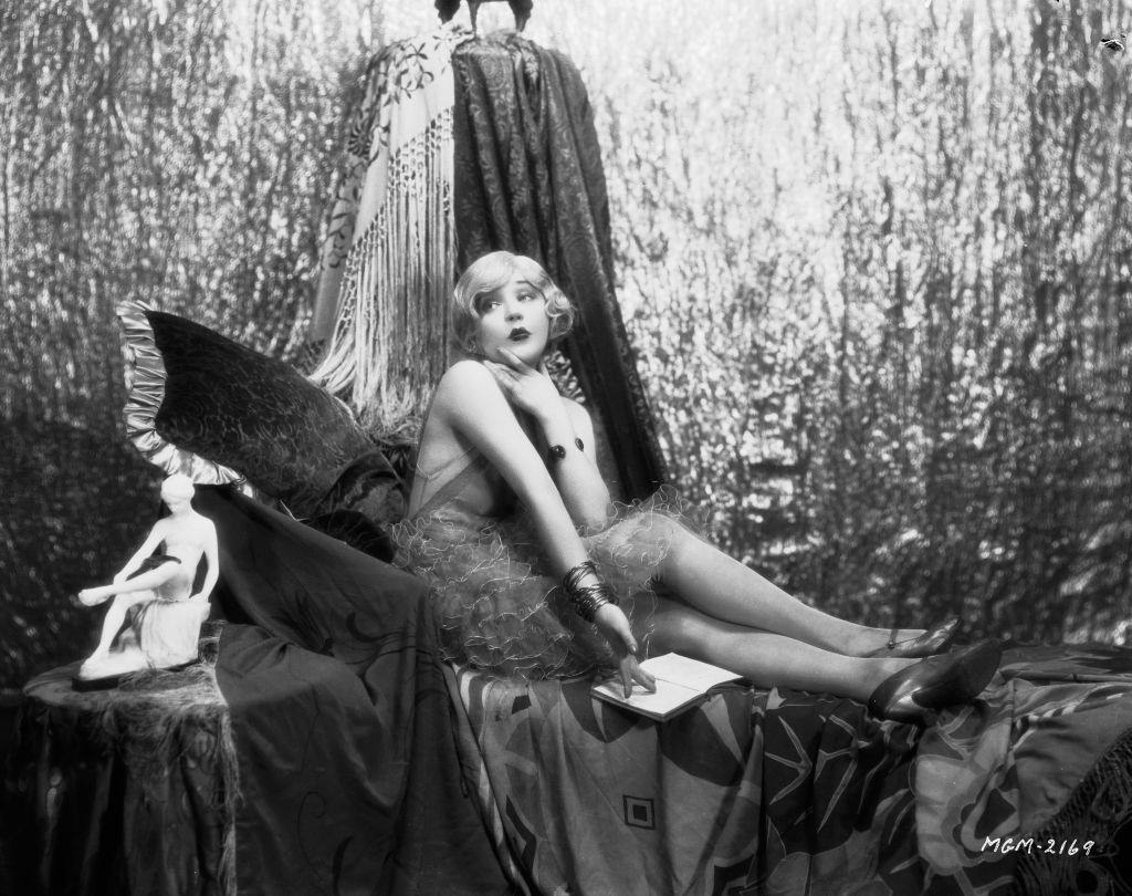 #44 Mae Murray poses with a book against a backdrop of draperies, 1926.