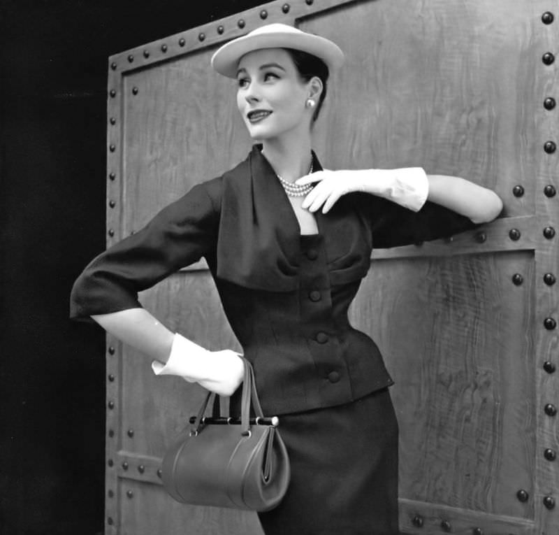 #40 Myrtle Crawford in navy blue wool afternoon suit, jacket has a large cowl neck held by a grosgrain buttoned placket, by Lanvin-Castillo, handbag by Violette Cornille, 1954