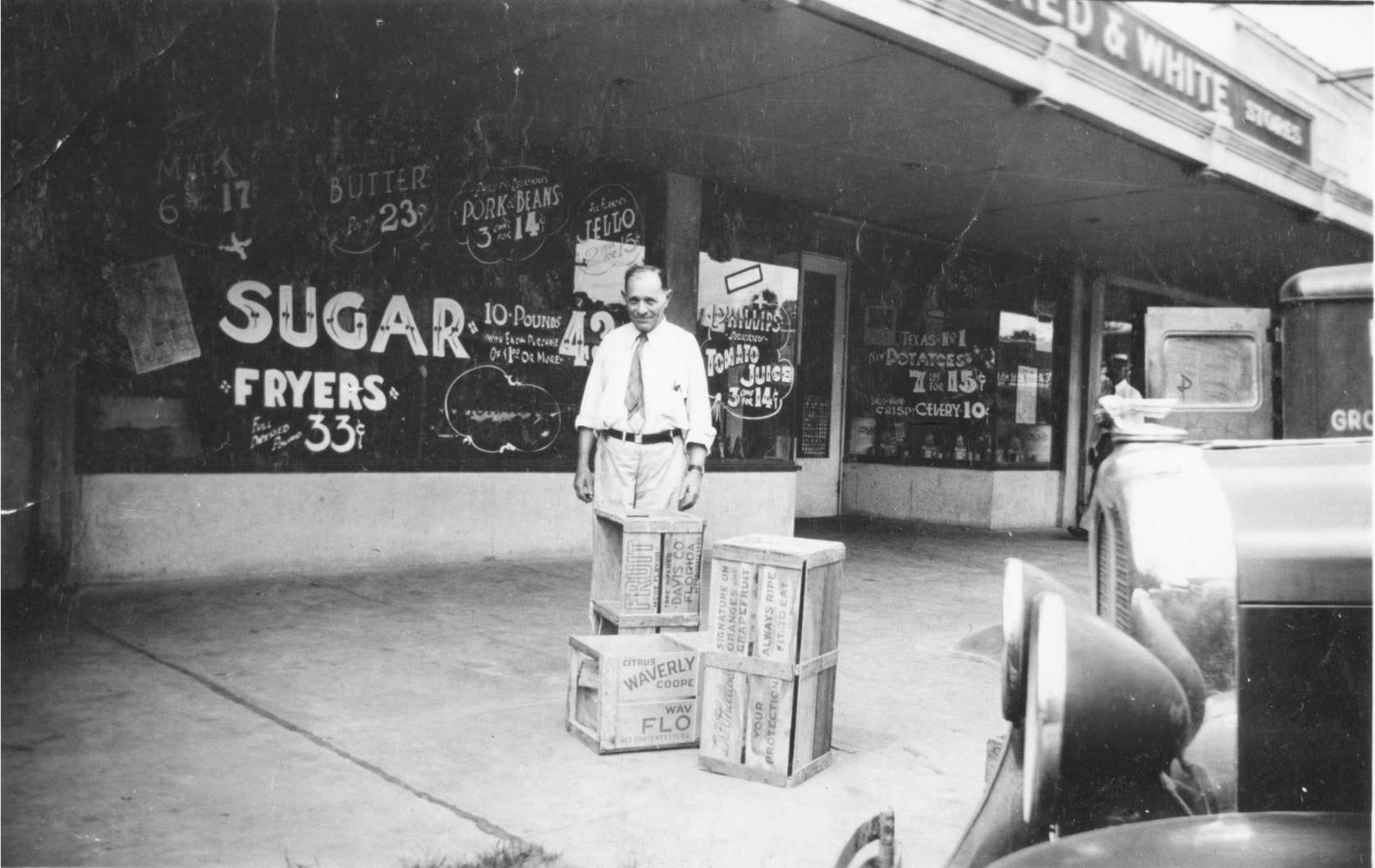 #47 Man in front of a store with wooden crates and car in foreground.
