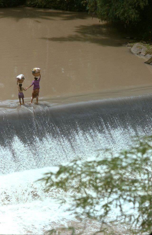 #54 A mother leads her young daughter by the hand across a levy that spans a stream inland from Tanah Lot, 1970s