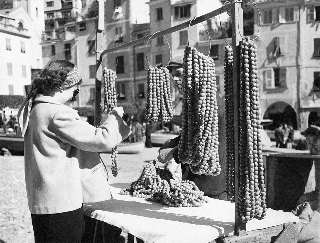 #28 A tourist takes a look on hazelnuts necklaces exposed in a stand, Portofino, 1960s.