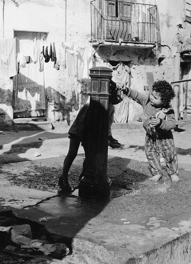 #42 A young child at a water fountain in a slum courtyard in Italy, 1960s.