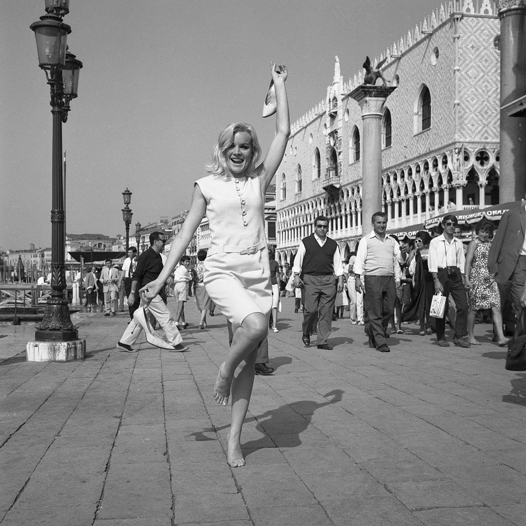 #45 American actress Carroll Baker, wearing a white dress in Riva degli Schiavoni while standing on her tiptoe, barefoot and holding her shoes, Venice, 1960s.