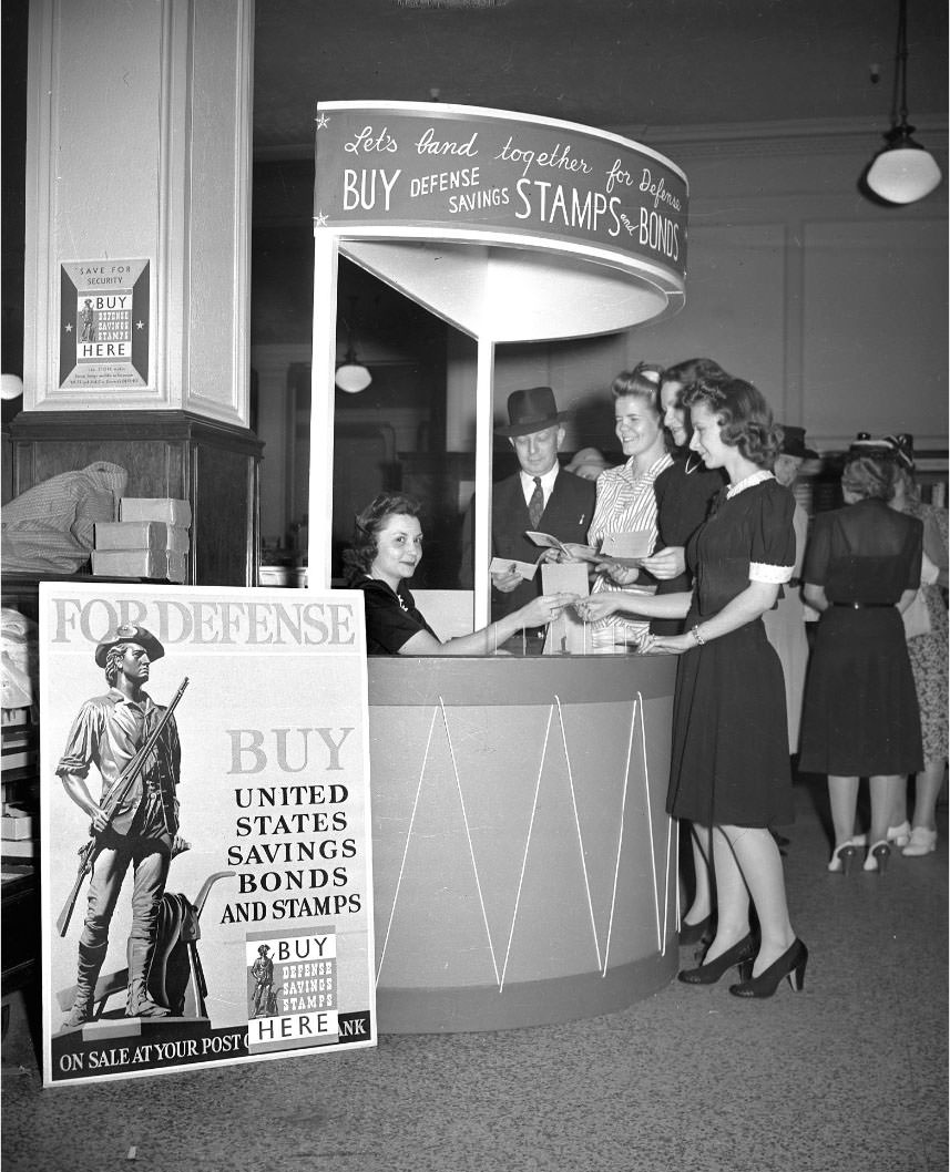 #2 A woman seated at a kiosk inside an unidentified building in Nashville, selling Defense Savings Stamps and Bonds.