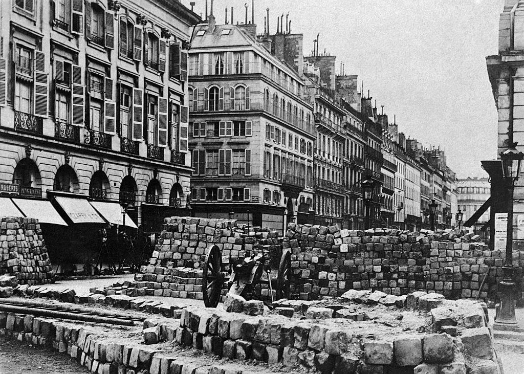 #21 A Paris street is barricaded with a stone-block wall and cannon, during the Franco-Prussian War.