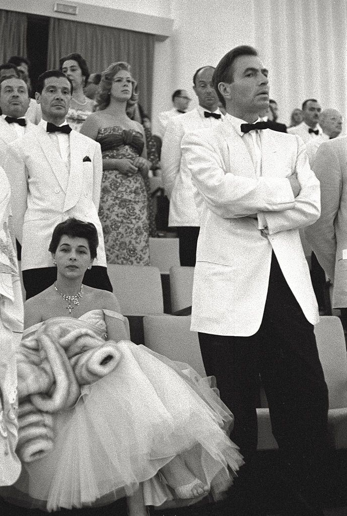 #149 James Mason waits standing with his arms crossed, his wife Pamela Kellino is seated next to him with an annoyed look.