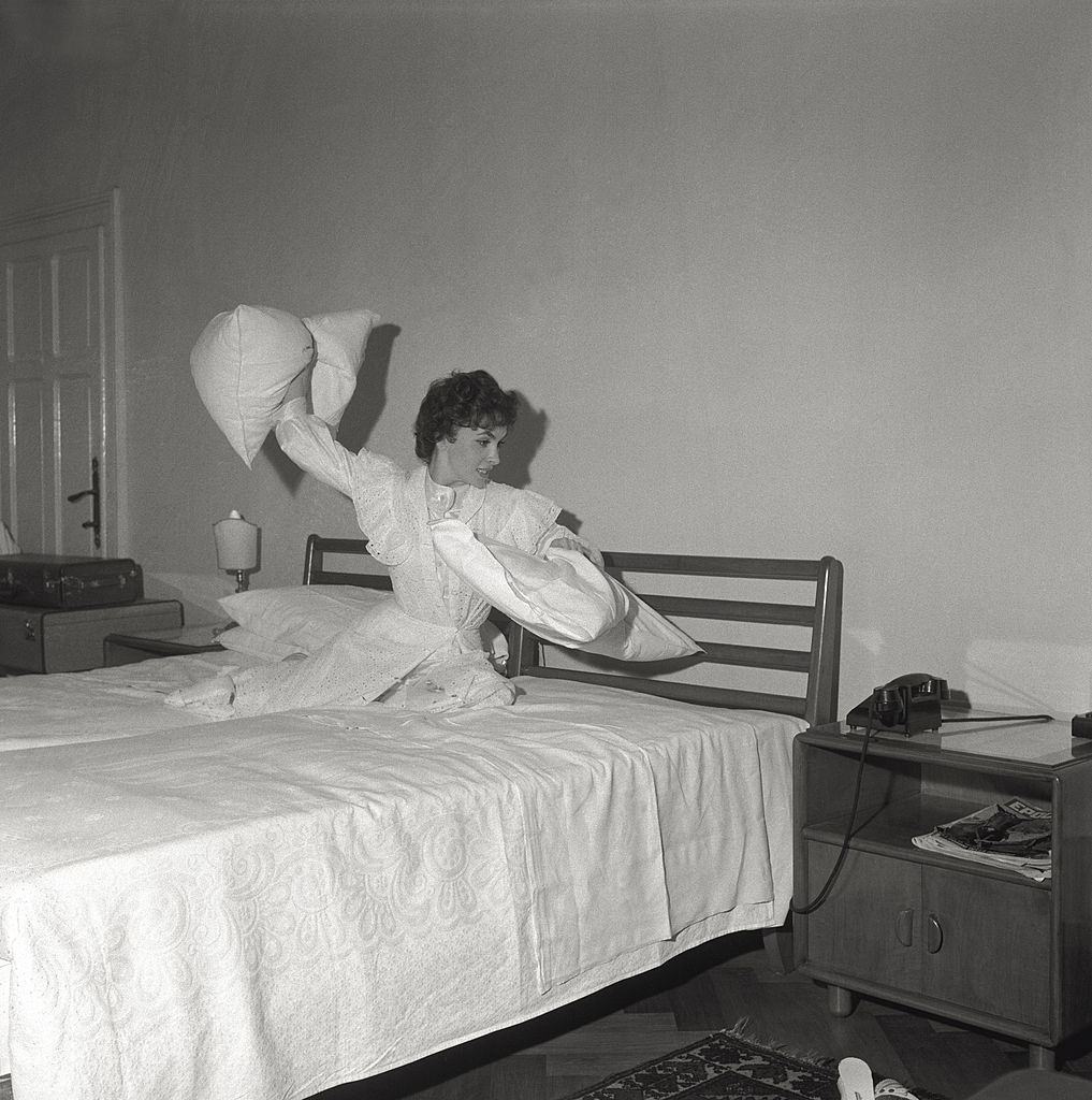 The actress Gina Lollobrigida plays with the pillows, sitting on the bed of a hotel room, during the 17th Venice Intenational Film Festival.