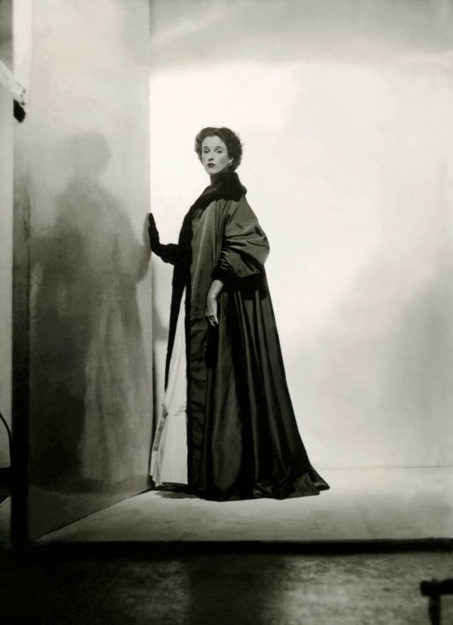 #30 Babe Paley standing with hand against a wall, and wearing a dark gray floor-length faille robe, 1946