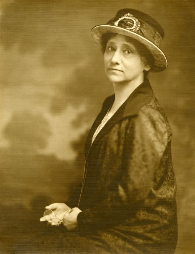 #25 Bertha Shambaugh wears a jacket and skirt with a floral design. Her hat has a matching decorative band, and she holds glasses, circa 1924