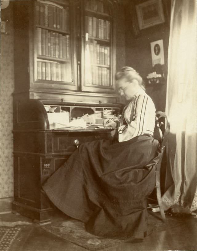 #10 Katherine Horack, pen in hand, seems to be writing at a desk in the sitting room, 1890s