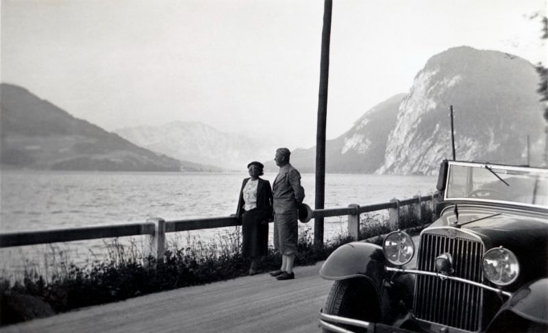 #2 A middle-aged couple posing with a Steyr XX Cabriolet on a gravel road running along the bank of a lake on a bleak summer’s day, 1930