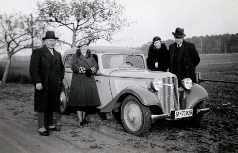 #13 Two stylish couples posing with a Adler Trumpf Junior Cabrio-Limousine on a dirt road in the countryside.