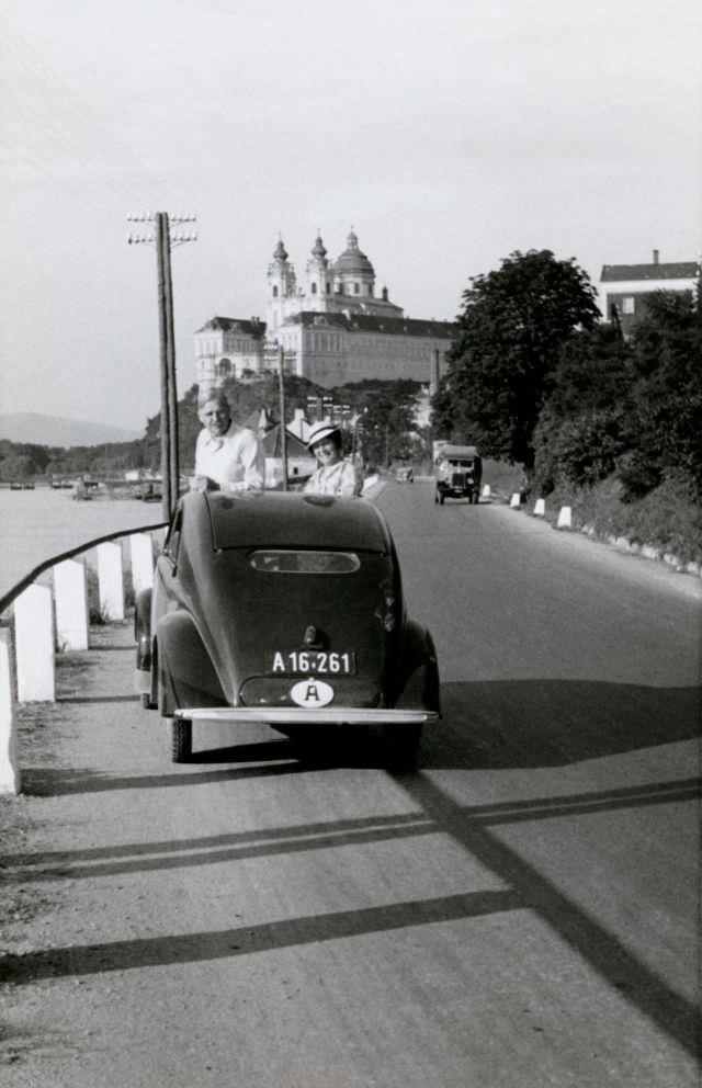 #19 A cheerful couple posing with a Steyr 50 on the side of the road in the town of Melk on the bank of the Danube with the famous monastery visible in the background.