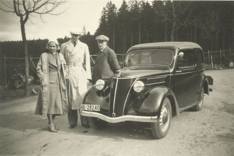 #28 A young elegant couple in trench coats and an elderly gent in a woollen coat posing with a German-built Ford Eifel.