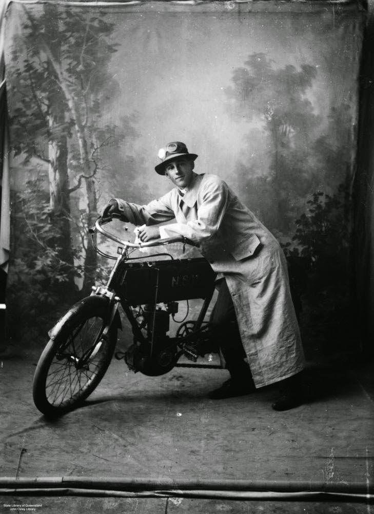 #6 Man dressed in a long dustcoat, hat and goggles with a motorcycle standing in front of a painted backdrop in a studio,1935.