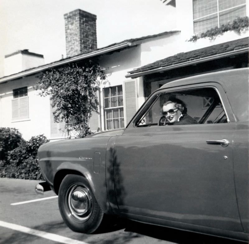 A blonde lady wearing dark sunglasses posing in the driver’s seat of a 1950 Studebaker Champion in front of a suburban home, 1950