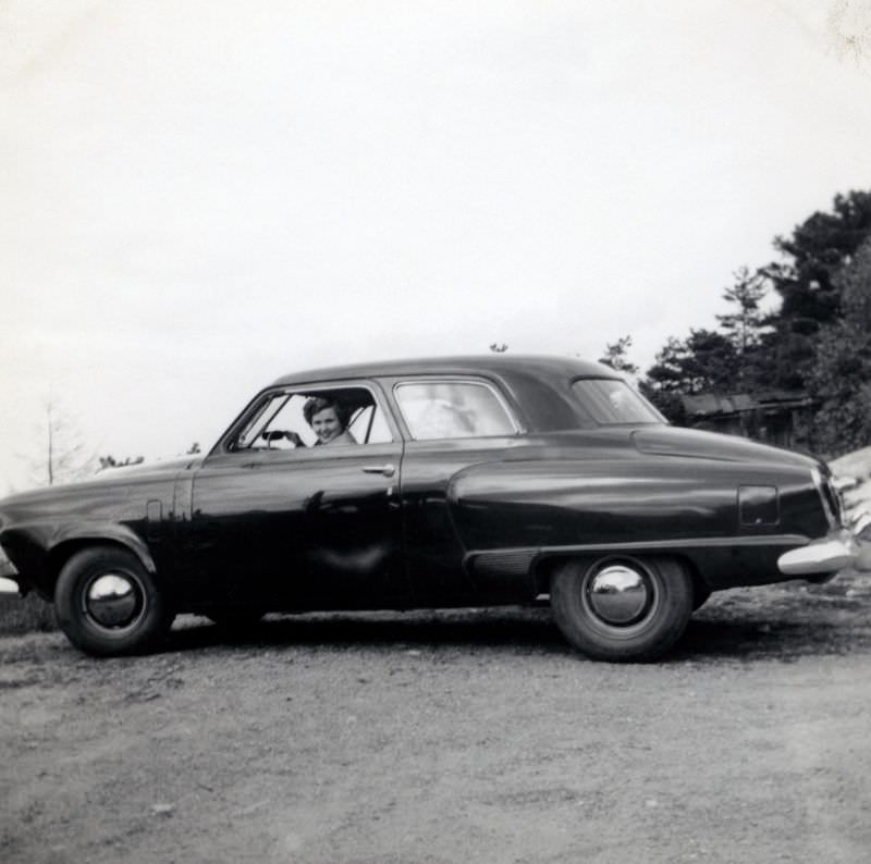 A cheerful lady posing in the driver’s seat of a 1950 Studebaker Champion Custom 2-Door Sedan in a gravel parking lot on the outskirts of town, 1950