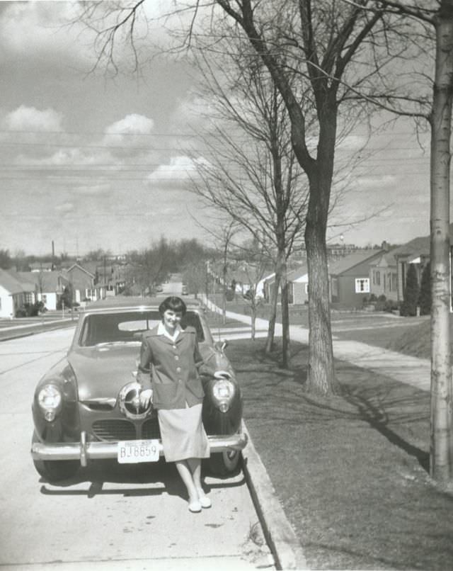 A young woman posing with a 1950 Studebaker Champion Convertible in a suburban street on a sunny winter’s day, 1952