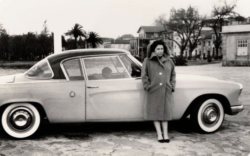 A slightly grumpy-looking lady in a woolen coat posing with a 1953 Studebaker Champion Regal Starliner on a bleak winter’s day.