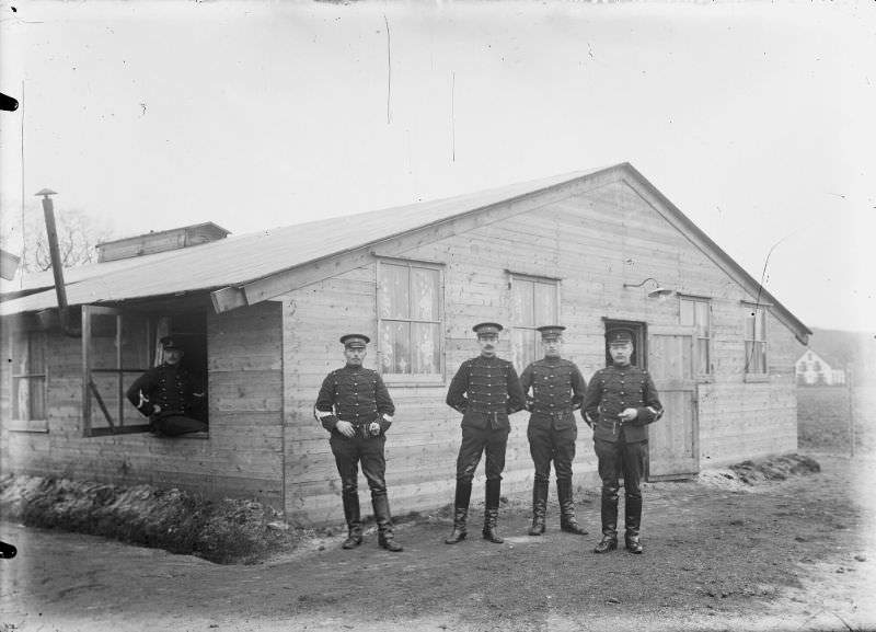 #24 Four NCOs standing in front of a barracks. A fifth NCO sits in a window of the barracks (left).