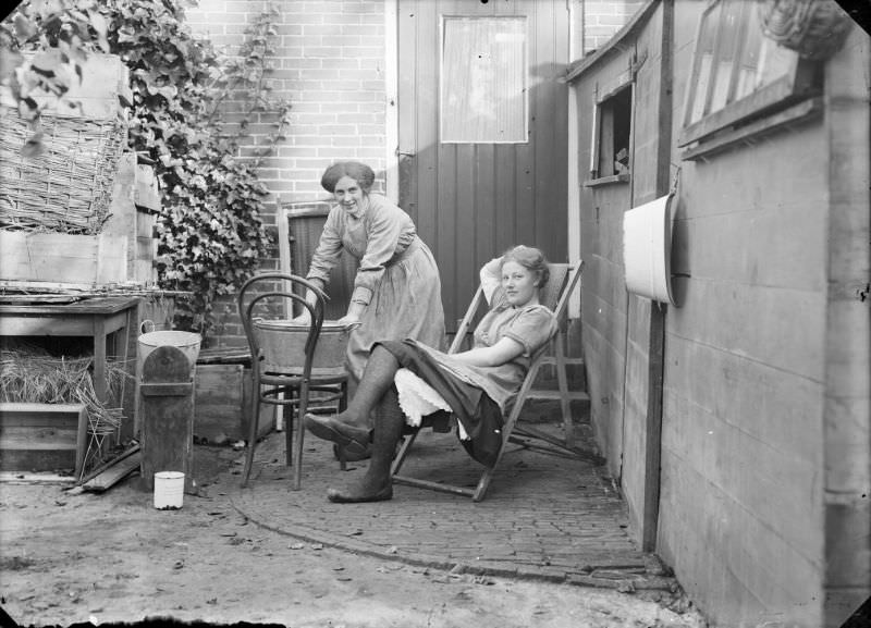 #28 Two women in a place behind a house. One of them is doing the laundry in a zinc tub, the other is lying in a deck chair, 1916