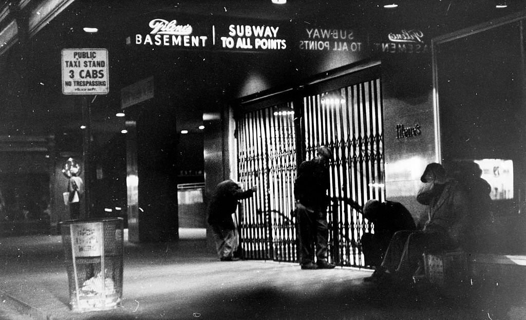 #10 Homeless people stand around a closed entrance to a subway station under Filene’s Basement in Downtown Crossing in Boston at night, September 1962.