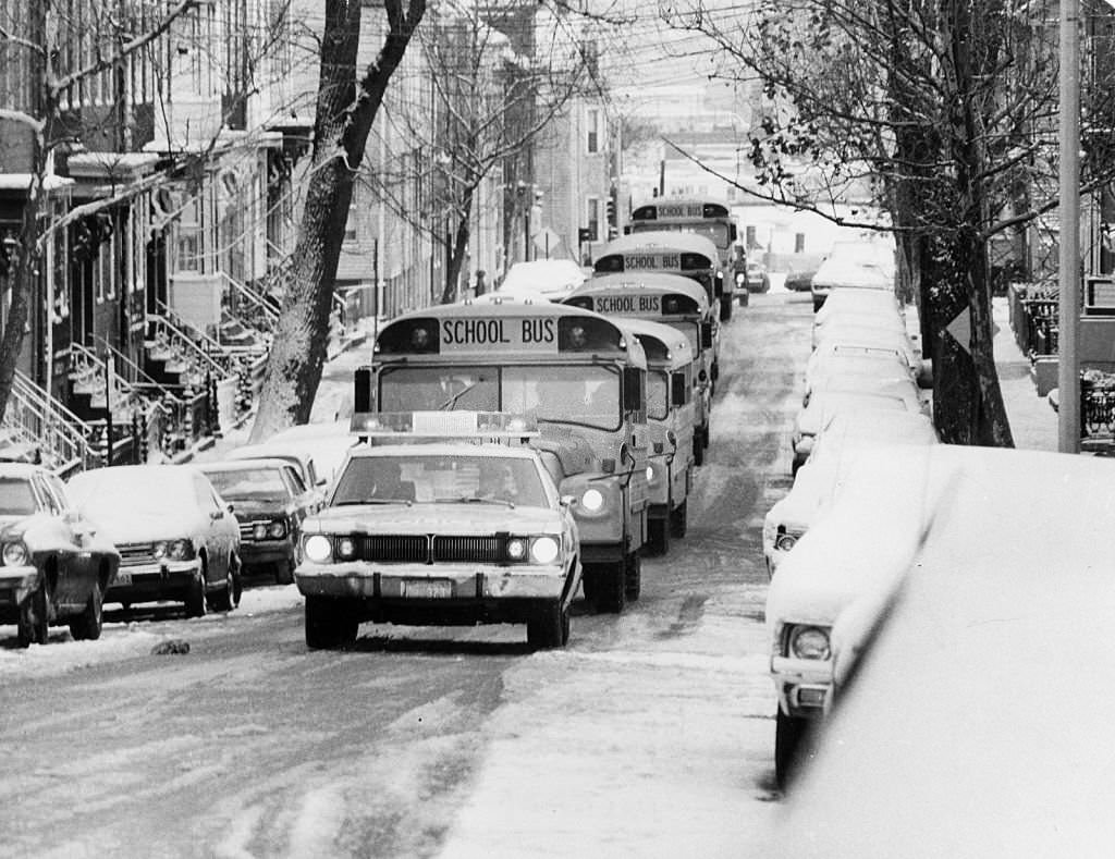 #138 On a snowy day, school buses follow a police car along G Street to South Boston High School in Boston, 1974.