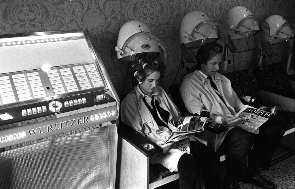 #8 A man and woman read magazines as they sit under hairdryers at the first unisex hairdressing salon in London, as music plays from a Wurlitzer jukebox, 1970