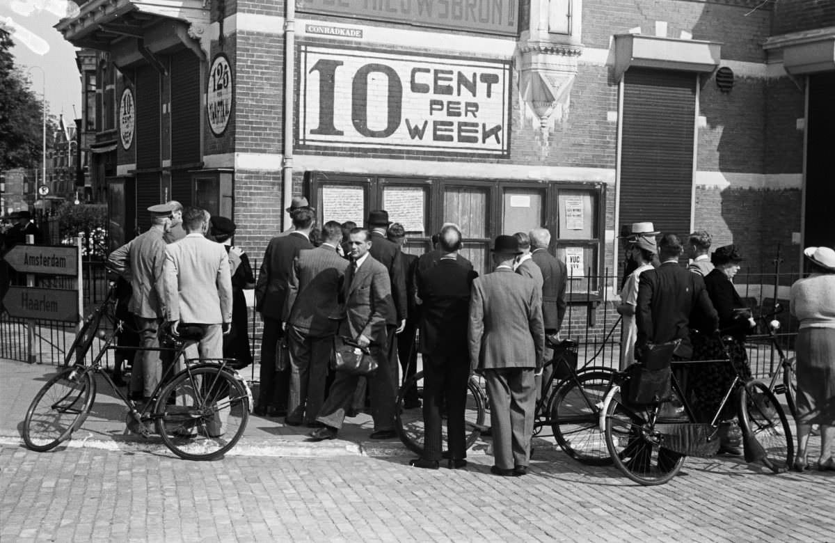 #9 A group of people read a series of handwritten wall papers in the street, under an advertisement for the newspaper “De Nieuwsbron”.