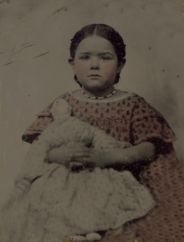 #8 A little girl in a pink dress holding a doll. Back of image has photographer card from C.B. Lawry, Farmington, Maine