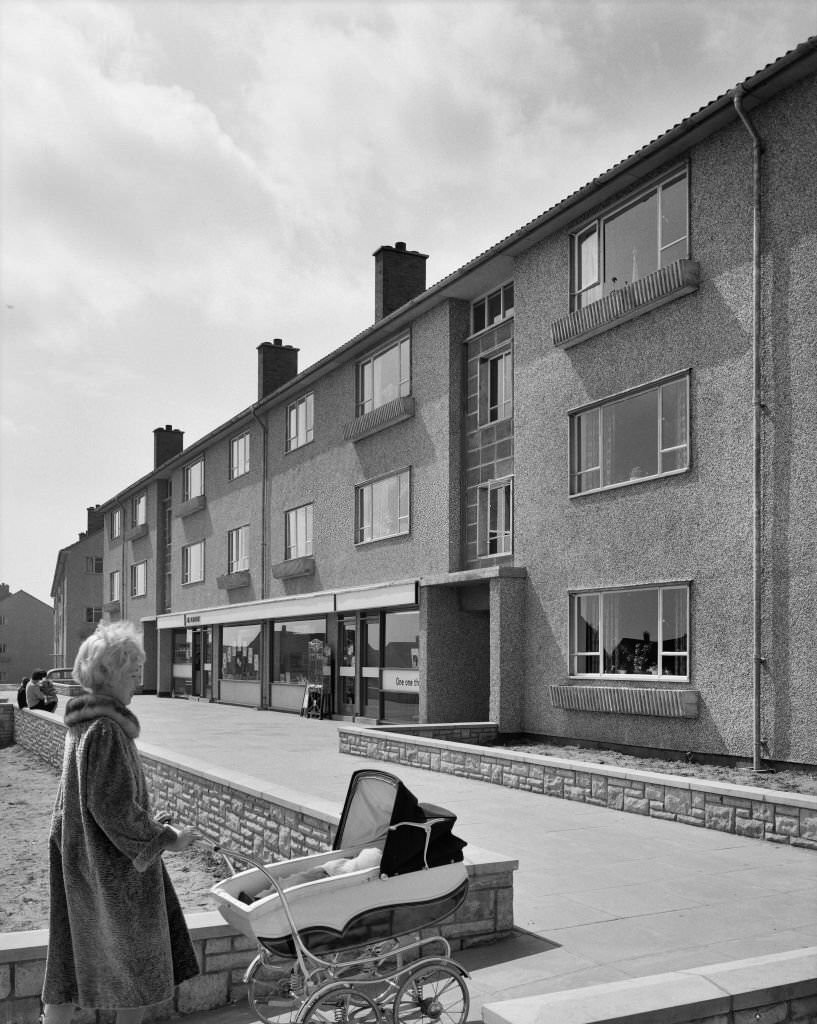 #64 A view of a block of Easiform flats with a ground floor shop in Chepstow, showing a woman pushing a pram in the foreground.