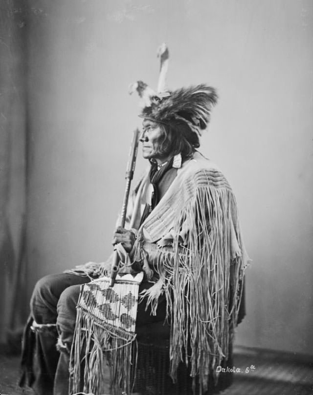 #15 Ta-Tan-Ka-Han-Ska (Long Fox or Long Buffal O Bull) in wearing feathered skunk hat and holding pipe and beaded and quilled bag.