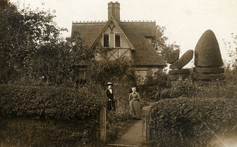 #43 Two women stand in the garden of a Victorian or Edwardian house built in a romantic cottage style.