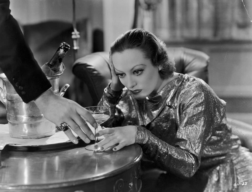 #45 Joan Crawford sits with her head in her hand as a man reaches to take her champagne glass, ‘Grand Hotel’.