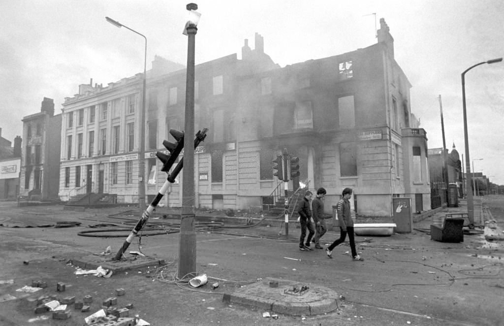 #115 Teenagers survey the aftermath of a second night of violent rioting in the Toxteth district of Liverpool, 1981