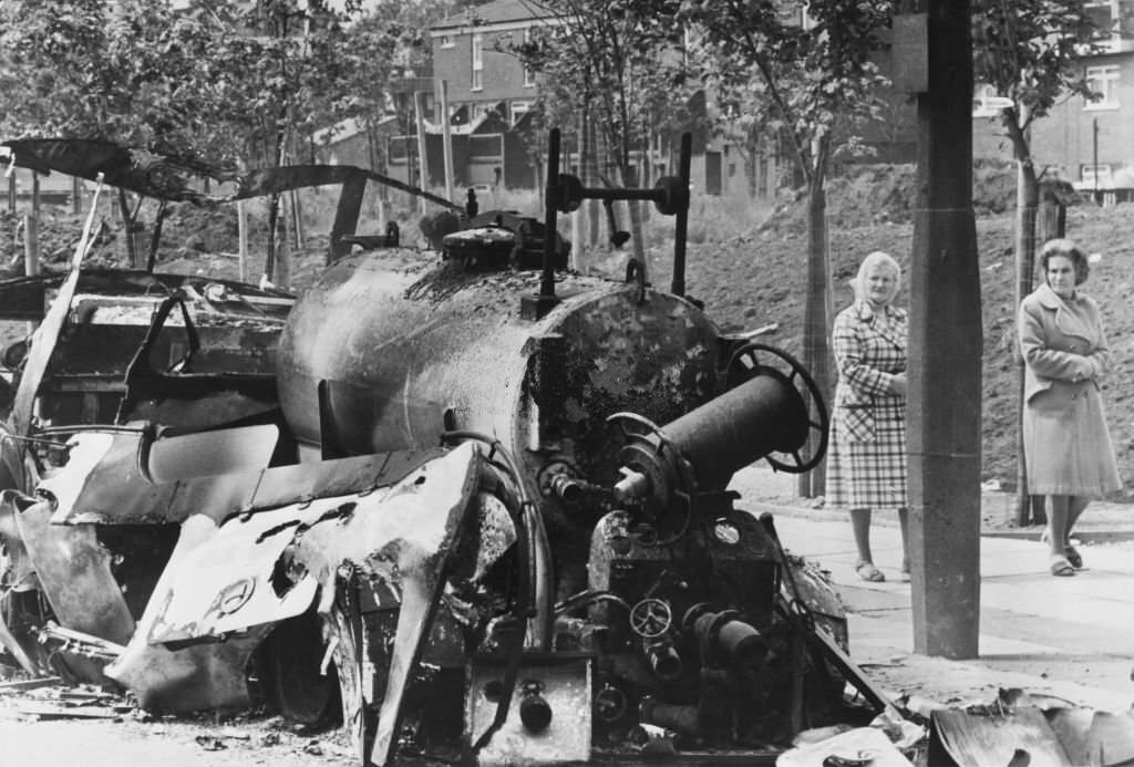 #63 Two women walk past the burnt wreck of a fire engine following a night of rioting in the Toxeth area of Liverpool, England, 7th July 1981.