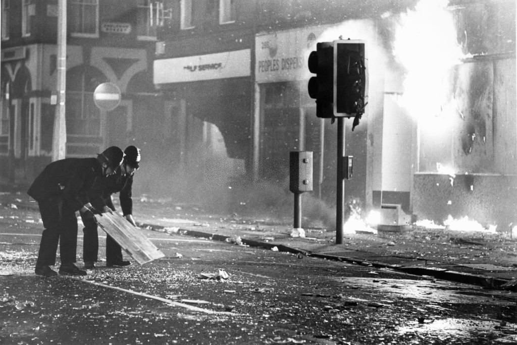 #107 Police officers try to clear broken glass from Park Road outside a burning looted shop, so that the fire brigade can control the fire.
