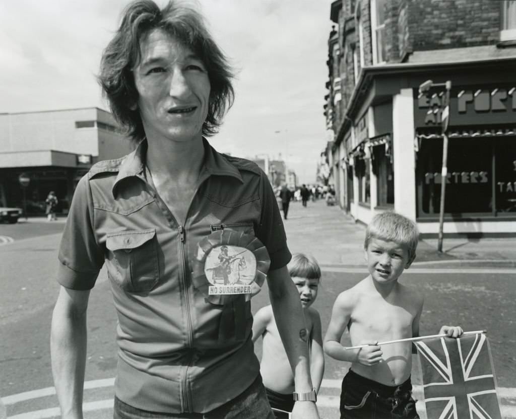 #156 A man wearing a rosette with the message ‘No Surrender’ accompanied by two young boys carrying a union jack flag on the streets of Liverpool, June 1982.