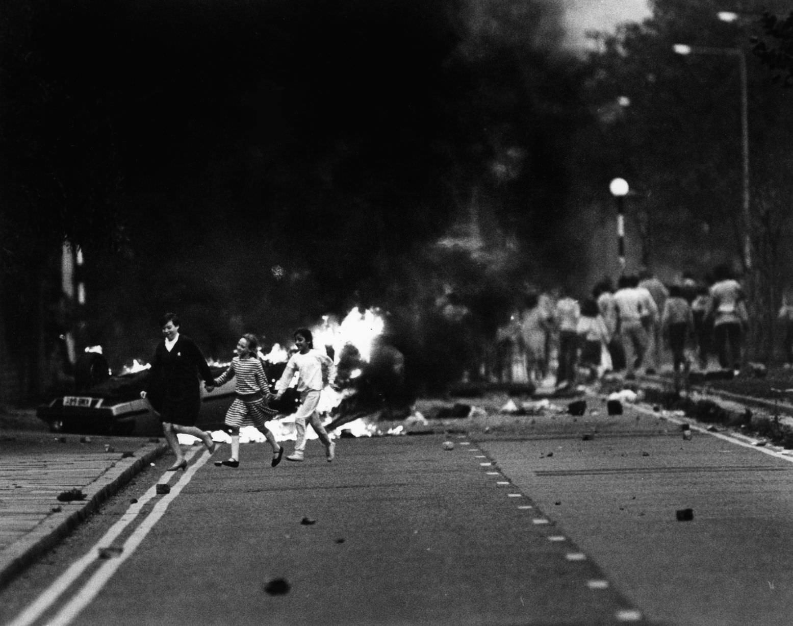 #51 Toxteth Riot 1st October 1985 Children crossing the street as the riot goes on in the background on Princes Road, Toxteth