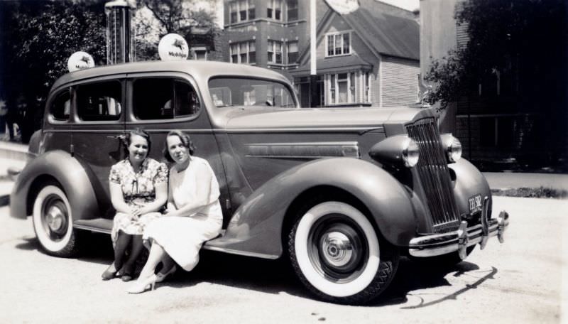 #13 Two cheerful young ladies posing on the running board of a 1936 Packard 120 Touring Sedan at a Mobile filling station in summertime.