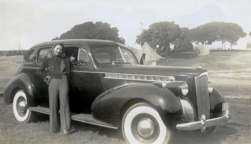 #17 A cheerful young lady in pants with a cigarette in her left hand posing with a 1940 Packard “120” Touring Sedan.