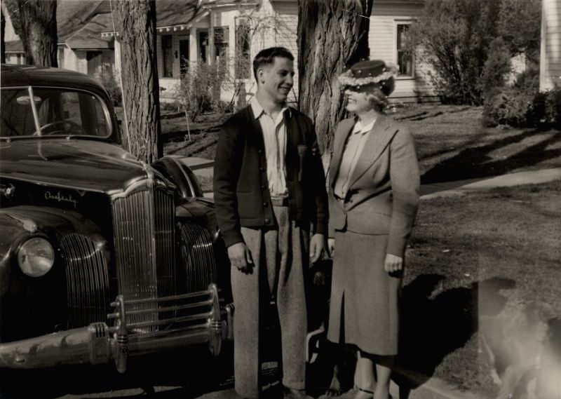 #19 A lady wearing a female suit and a fancy hat and a man in a woolen cardigan posing in front of a 1941 Packard One Sixty, 1946