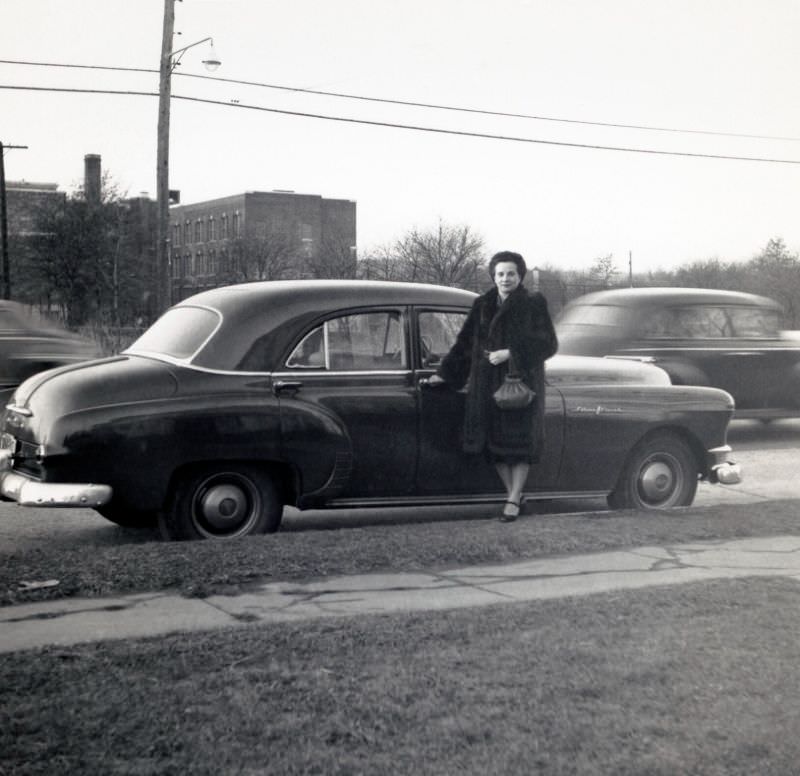 A middle-aged lady in a fur coat posing at the passenger side door of a 1950 Pontiac Chieftain Sedan on a bleak winter’s day, 1952
