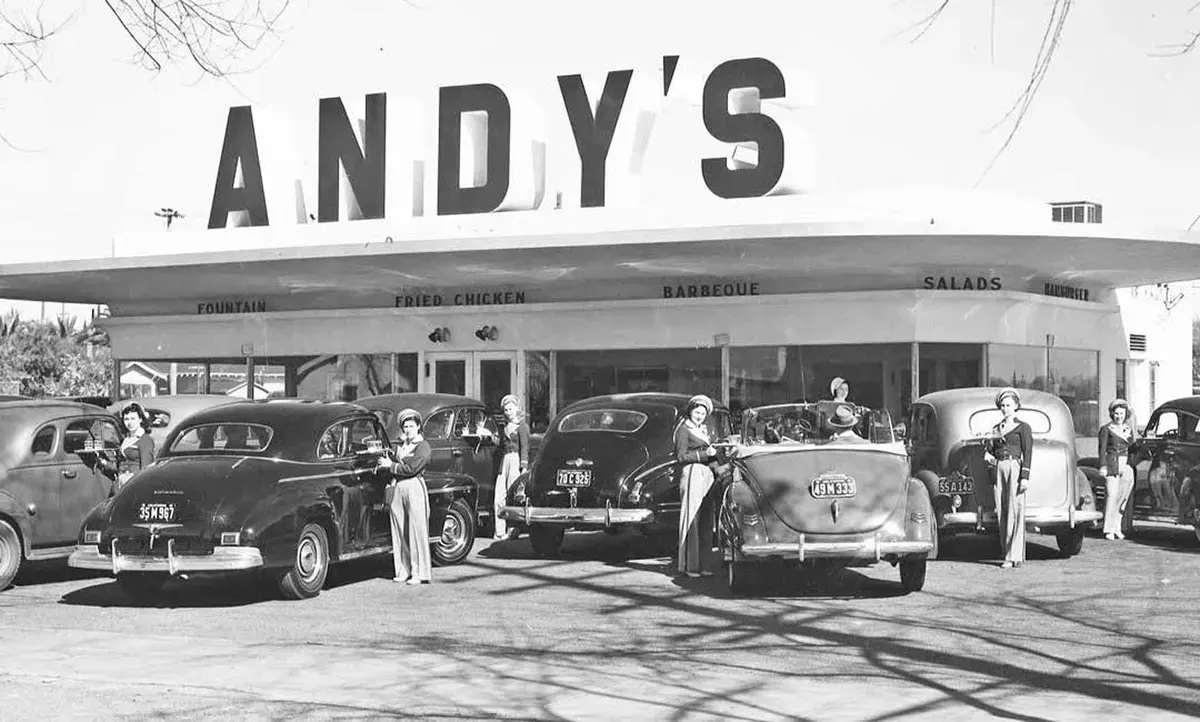 #44 Andy’s Drive-In with carhops pose in front with cars they’re serving, 1941.