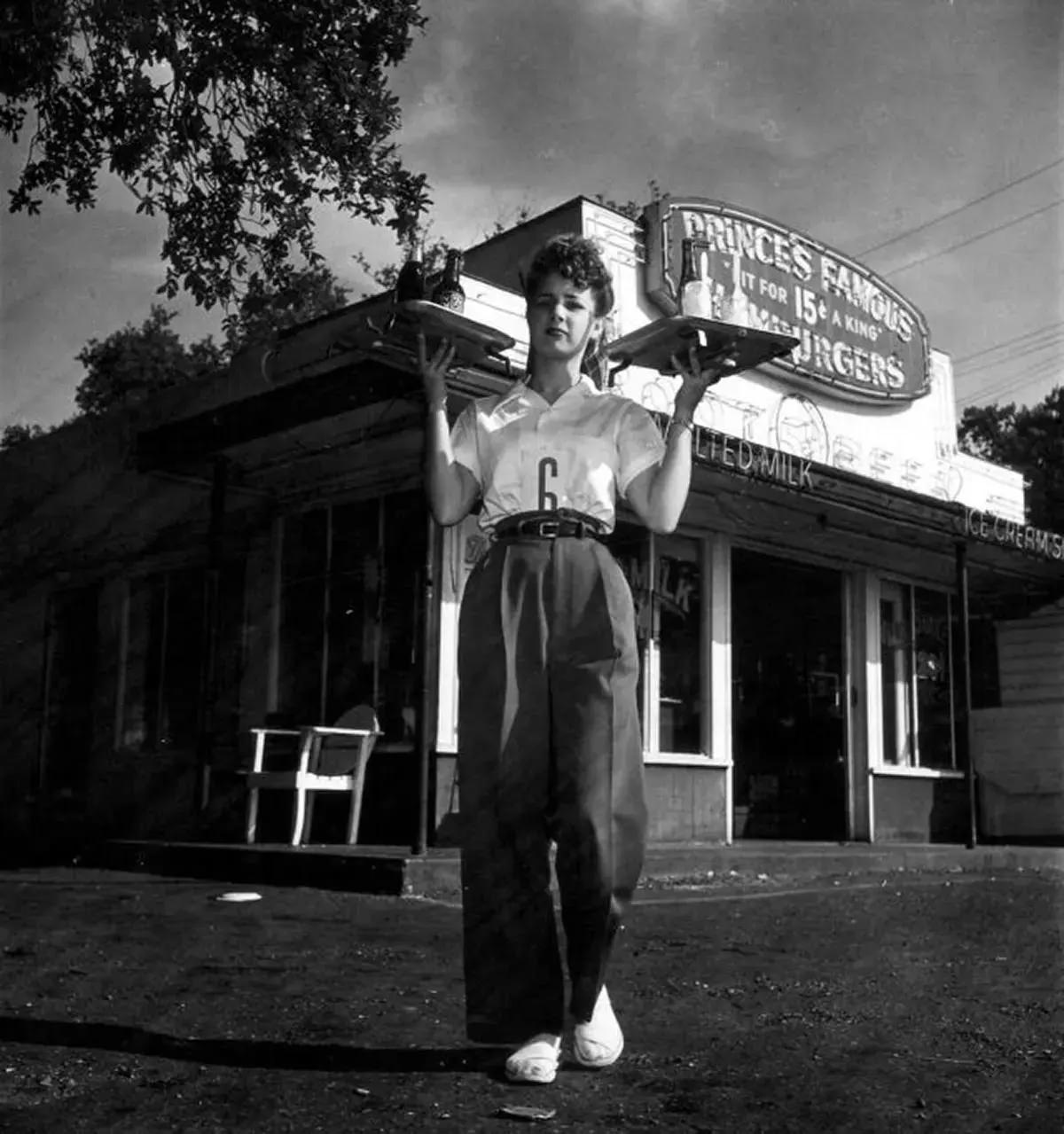 #46 A carhop holds two trays as she poses for a shot in front of a small diner, Houston, Texas, 1945.