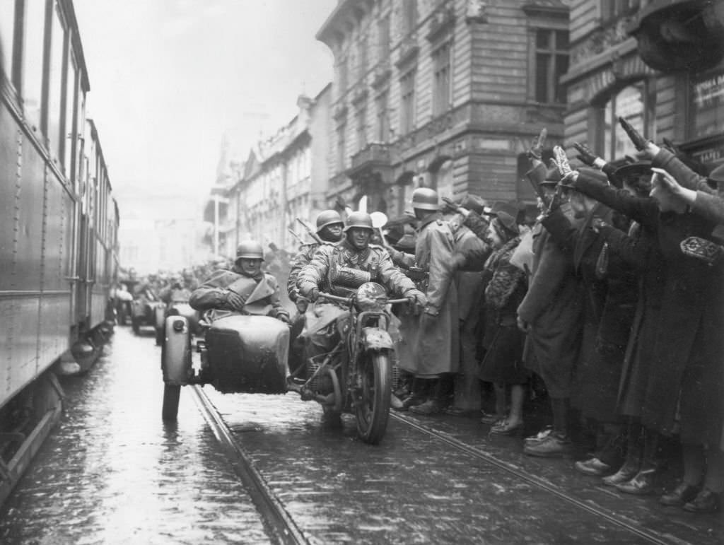#46 Crowds on the sidewalk salute Nazi soldiers riding in a motorcycle infantry unit as German troops enter Prague, Czechoslovakia.